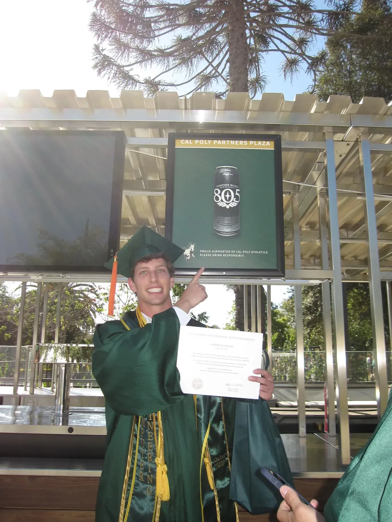 Luke in Cal Poly cap and gown, holding his commencement certificate and pointing at a Firestone Walker 805 sign overhead